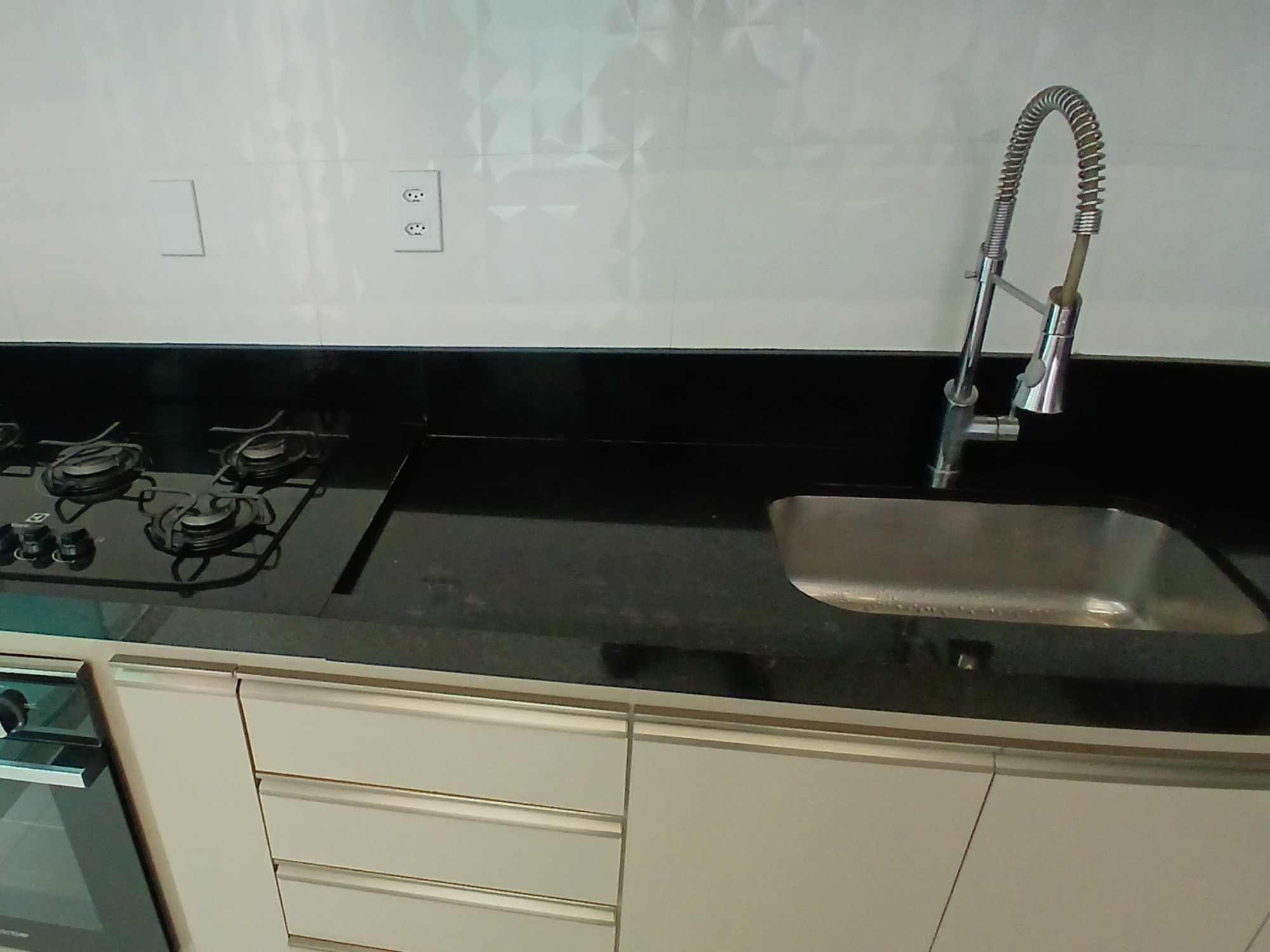 Bancada de granito preto com cooktop, torneira gourmet e cuba de inox, sobre armários claros e forno embutido.