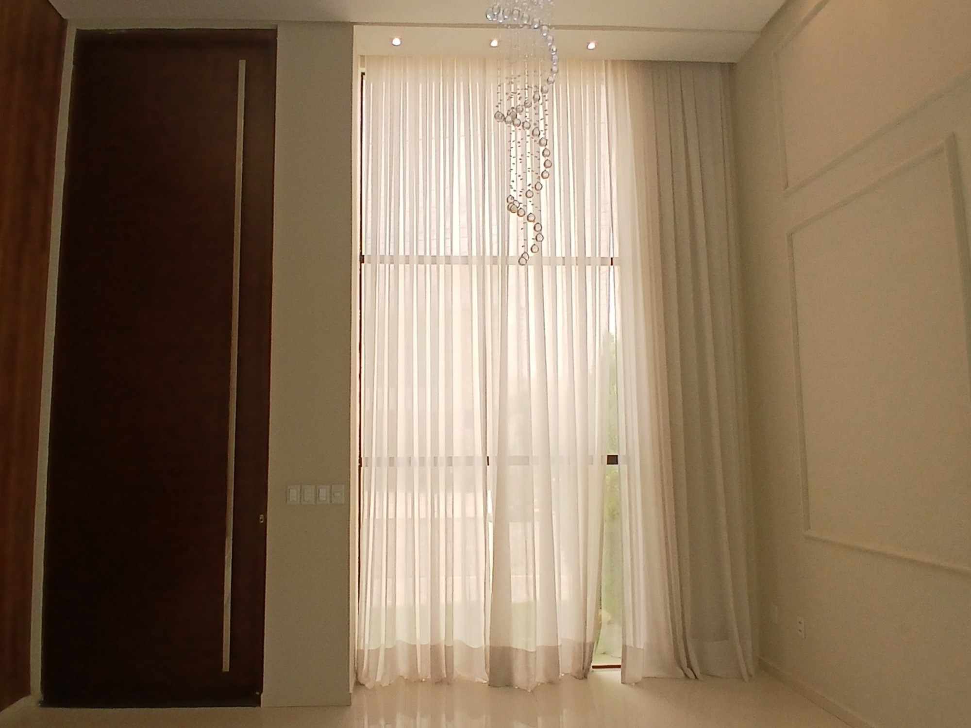 Sala de pé-direito duplo com cortina do piso ao teto, porta de madeira e lustre de cristal.