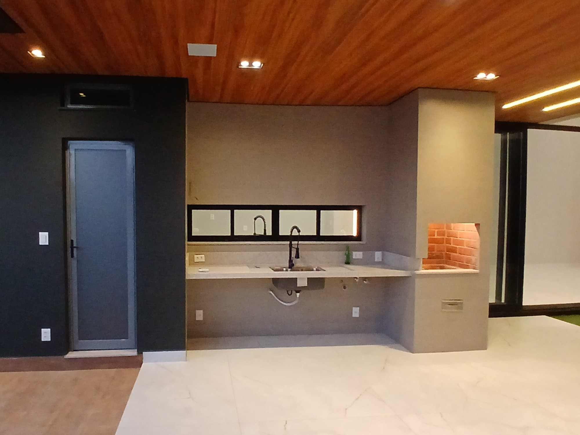 Área gourmet com forro de madeira, perfis de LED, bancada e churrasqueira revestida de tijolo.