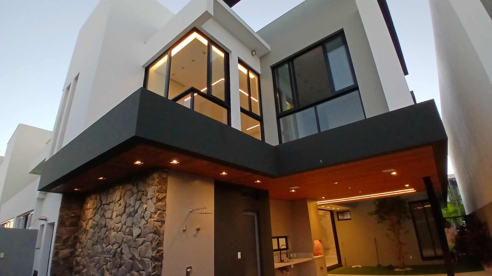 Fundos da casa nova ao entardecer, com parede de pedra, iluminação embutida e área gourmet.