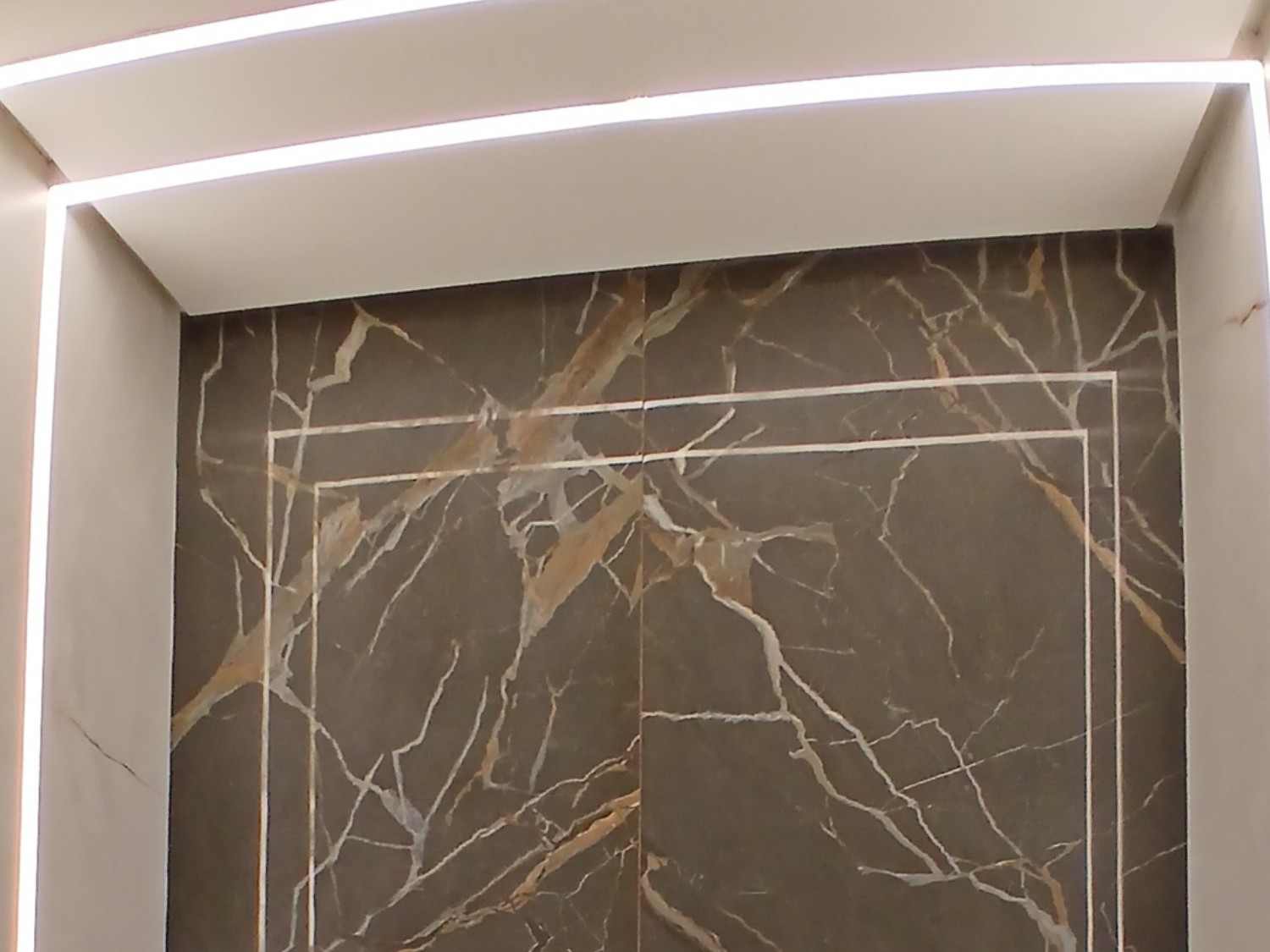 Parede de porcelanato marmorizado escuro com moldura de metal e perfis de LED no teto do lavabo.